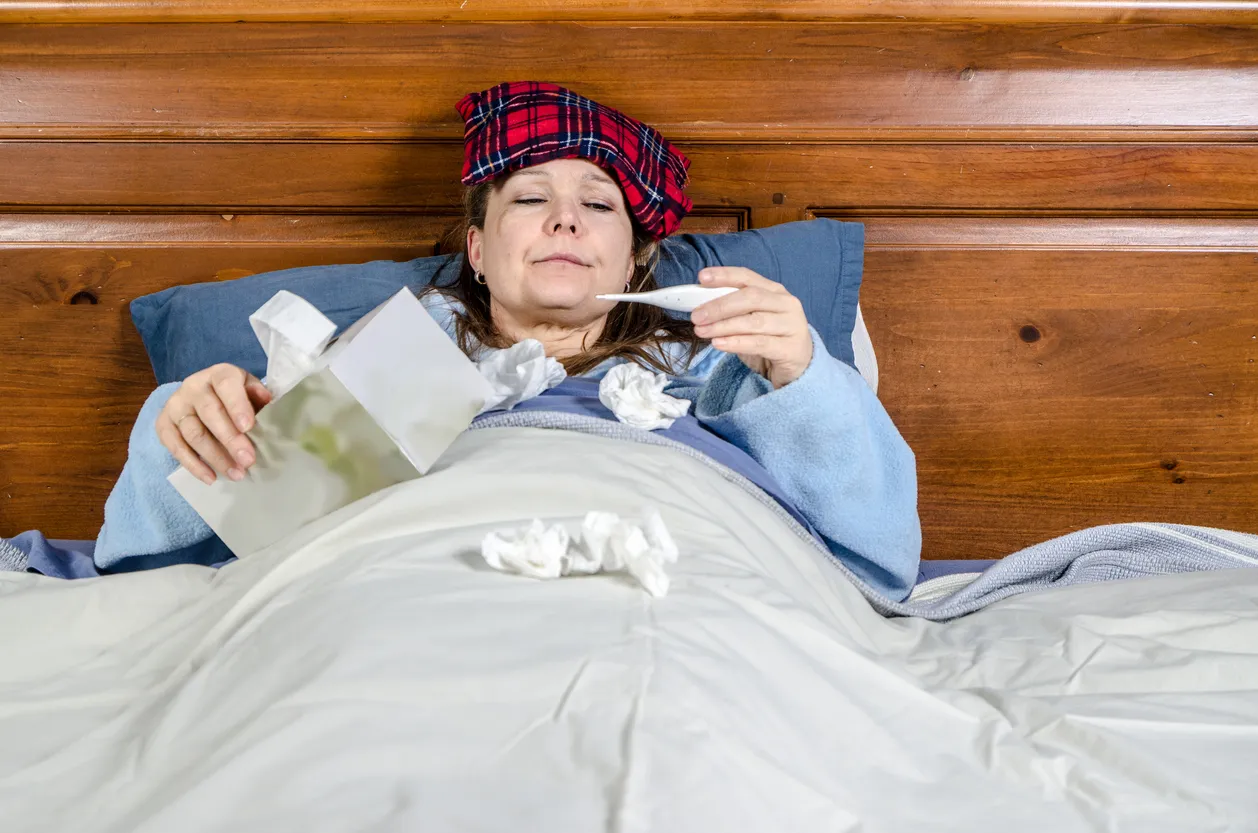 Femme malade au lit avec une boîte de mouchoirs et regardant un thermomètre @istockphoto/Marc Dufresne